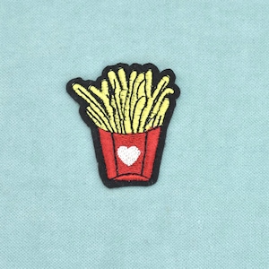 Könnte beinhalten: Ein gestickter Aufnäher, der eine rote Schachtel mit gelben Pommes frites zeigt. Die rote Schachtel hat ein weißes Herz in der Mitte auf der Vorderseite. Das gesamte Design ist mit einem dicken schwarzen Rand versehen, der eine klare Form bildet.