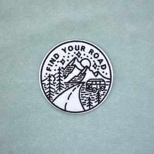 Peut inclure: Un écusson rond noir et blanc avec le texte "FIND YOUR ROAD" encerclant un paysage de montagne. Le motif comprend une route, des arbres, un van, des étoiles et un soleil. L'écusson est sur un tissu vert clair.