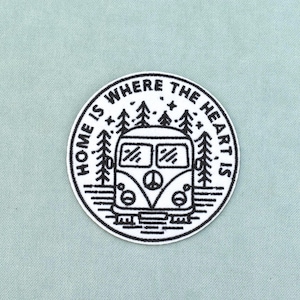 Può includere: Una toppa ricamata circolare con un disegno a linee bianche e nere di un camper con un simbolo della pace sulla griglia anteriore, circondato da pini e stelle, con il testo "HOME IS WHERE THE HEART IS" arcuato lungo la parte superiore.