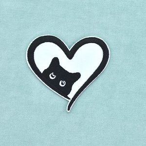 May include: A black and white embroidered patch featuring a heart shape with a thick black border. Inside the white heart, a black cat head is visible, peeking out from the lower left side with two white eyes looking forward.