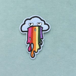 Può includere: Toppa ricamata raffigurante una nuvola azzurra con contorno nero e un design a vomito arcobaleno. La toppa ha uno stile stravagante e fumettistico ed è ambientata su uno sfondo di tessuto azzurro.
