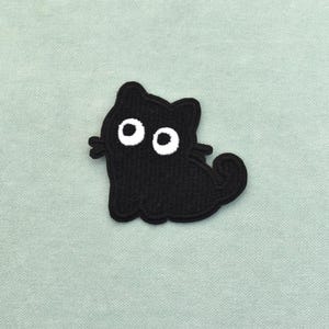 Op de afbeelding: Een zwarte kattenpatch met witte ogen en snorharen. De kat zit met opgerolde staart. De patch is gemaakt van een textuurrijk materiaal tegen een lichtgroene achtergrond.