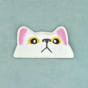 Op de afbeelding: Een geborduurde strijkapplicatie in de vorm van een witte kattenkop met roze binnenoren, grote gele ogen en een donkergrijze neus en mond. De kattenkop heeft een platte onderkant en puntige oren, uitgevoerd in wit borduurgaren.