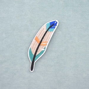 Peut inclure: Écusson de plume brodée avec une bordure blanche. La plume présente un corps beige avec des accents turquoise, orange et bleu, et une tige noire. L'écusson est sur un fond de tissu bleu clair.
