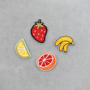 Pode incluir: Remendos de frutas bordadas, incluindo um morango vermelho, um cacho de bananas amarelas, uma fatia de limão e uma fatia de laranja. Cada remendo tem uma borda preta e está sobre um fundo cinza.