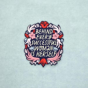 Può includere: Una toppa ricamata blu scuro con il testo BEHIND EVERY SUCCESSFUL WOMAN IS HERSELF in lettere color crema, circondata da un bordo decorativo di fiori e foglie rosa, rossi e bianchi su uno sfondo di tessuto azzurro.