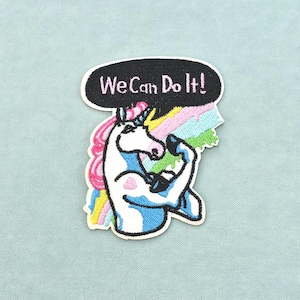 Peut inclure: Un écusson brodé représentant une licorne blanche avec une crinière rose et une corne bleue, fléchissant son bras dans une pose de biceps. Derrière la licorne se trouve un arc-en-ciel, surmonté d'une bulle noire avec le texte blanc "We Can Do It!".
