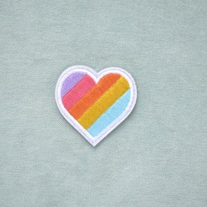Peut inclure: Écusson brodé en forme de cœur avec un motif arc-en-ciel. Le patch a une bordure blanche et un cœur rempli de rayures diagonales violettes, roses, oranges, jaunes et bleu clair. Le patch est sur un tissu vert d'eau clair.