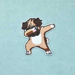 Könnte beinhalten: Ein gestickter Aufnäher, der einen beigen und braunen Mops in einer Dab-Pose zeigt. Der Mops hat ein dunkelbraunes Gesicht, einen weißen Brustfleck und ist so dargestellt, dass der Kopf in der Armbeuge eines erhobenen Arms liegt.