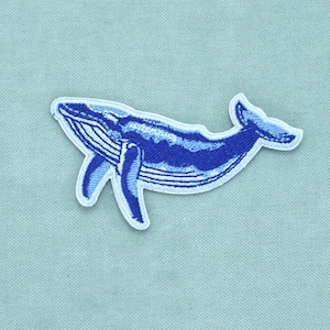 Peut inclure: Un écusson brodé représentant une baleine à bosse dans des tons bleu foncé et bleu clair. La baleine est représentée en position de nage avec ses nageoires pectorales étendues et sa nageoire caudale visible, bordée d'un contour blanc.