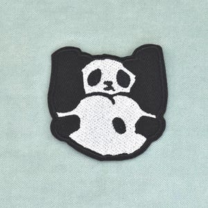 以下が含まれることがあります： 白と黒のパンダをデザインした刺繍ワッペン。白い顔、黒い目の周りの模様、白い体に黒い円形の斑点がある胸部が特徴で、全体が太い黒い縁取りで囲まれています。