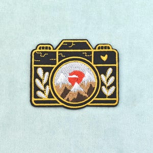May include: An embroidered patch shaped like a camera with a black body and yellow outlines. The lens features a circular scene of brown mountains with white snow-capped peaks under a bright red sun. White leaf sprigs flank the lens.