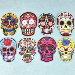 May include: Eight embroidered skull patches arranged in two rows. Each patch features intricate, colourful designs including floral patterns, crosses, and geometric shapes in vibrant hues of yellow, red, black, white, blue, and pink on fabric.