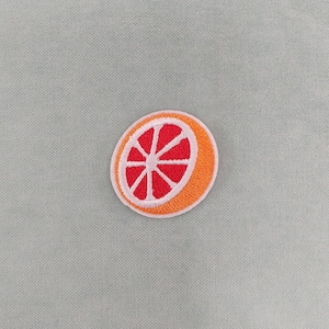 May include: Embroidered patch featuring a grapefruit slice design. The patch has a bright orange rind, a white border, and a vibrant red interior with white segments. The patch is circular and likely for clothing or accessories.