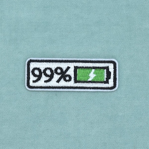 Op de afbeelding: Een rechthoekige geborduurde patch met een witte achtergrond en een zwarte rand. Het ontwerp toont de tekst 99% in het zwart naast een groen batterijpictogram met een wit bliksemschichtsymbool, wat een bijna volle lading vertegenwoordigt.