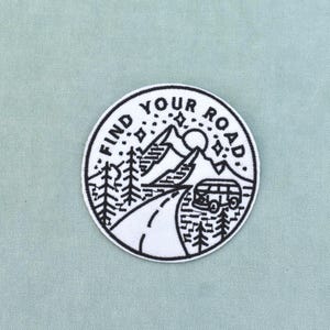 Pode incluir: Um emblema bordado circular com fundo branco e costuras pretas. O design retrata uma estrada sinuosa que leva a montanhas, pinheiros, uma carrinha de campismo e estrelas, com o texto FIND YOUR ROAD arqueado no topo.