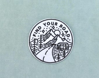 May include: A circular embroidered patch featuring a white background with black stitching. The design depicts a winding road leading toward mountains, pine trees, a camper van, and stars, with the text FIND YOUR ROAD arched at the top.