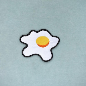 Peut inclure: Écusson brodé en forme d'œuf au plat. Le blanc d'œuf est blanc avec un bord noir, et le jaune est jaune avec un centre orange. L'écusson est sur un fond bleu clair.