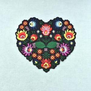 Peut inclure: Un écusson en tissu en forme de cœur avec une broderie complexe sur fond noir. Le motif présente une disposition symétrique de fleurs colorées dans des tons de rouge, jaune, rose et bleu, agrémentées de feuilles et de tiges vertes.