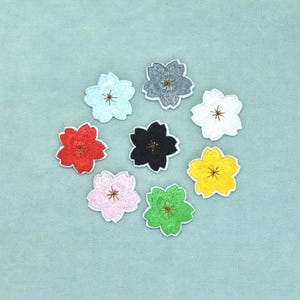 Pode incluir: Uma coleção de oito emblemas bordados em forma de flor dispostos num padrão circular. Os emblemas apresentam flores de cinco pétalas em várias cores, incluindo azul claro, cinzento, branco, vermelho, preto, amarelo, verde e rosa claro com centros castanhos.