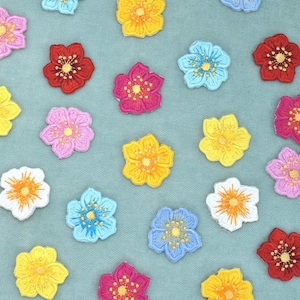 Pode incluir: Uma coleção de pequenos remendos de flores de cinco pétalas em várias cores, incluindo vermelho, amarelo, azul, rosa e branco. Cada remendo bordado apresenta um centro detalhado com fios radiantes, espalhados sobre uma superfície azul-clara.