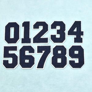 May include: A set of ten iron-on patches featuring the numbers zero through nine in a dark navy blue colour. Each number has a bold, block-style font with a clean white border outline, arranged in two rows of five against a light blue background.
