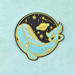 Peut inclure: Un écusson brodé circulaire représentant une baleine bleu clair nageant dans un espace noir. Le motif comprend une planète jaune avec des anneaux, plusieurs étoiles jaunes et une queue de baleine bleu clair dépassant du bord du cercle.