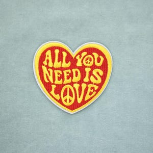 Peut inclure: Écusson brodé en forme de cœur avec l'inscription "ALL YOU NEED IS LOVE" en jaune, orné d'un symbole de paix. Le patch a un fond rouge avec une bordure jaune, sur un tissu bleu clair.