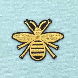 May include: An embroidered patch featuring a stylised bee design. The bee is rendered in metallic gold thread with black thread detailing for the body segments and outline, set against a solid black background shape that follows the insect's form.