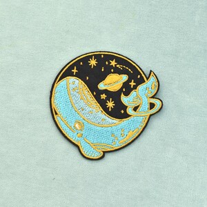 Op de afbeelding: Een ronde geborduurde patch met een lichtblauwe walvis die door een zwarte ruimteachtergrond zwemt. Het ontwerp bevat een gele planeet met ringen, verschillende gele sterren en een vallende ster. De walvis heeft een gebogen staart.