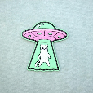 Può includere: Toppa ricamata raffigurante un gatto bianco rapito da un UFO rosa e verde. La toppa ha un bordo nero e si trova su uno sfondo azzurro. Il gatto ha un'espressione sorpresa.