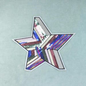 Könnte beinhalten: Ein fünfzackiger Sternaufnäher mit einem mehrfarbigen Paillettenmuster. Die Oberfläche besteht aus diagonalen Streifen aus Pailletten in Blau-, Rosa-, Silber-, Grün- und Violetttönen, die von einem dünnen weißen Stoffrand und schwarzen Nähten umgeben sind.