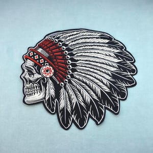 Peut inclure: Écusson brodé représentant un crâne portant une coiffe amérindienne. La coiffe est rouge et blanche, avec des plumes noires et blanches. Le crâne est blanc avec des ombres noires, et l'écusson a une bordure noire.
