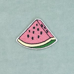 Può includere: Una toppa ricamata triangolare a forma di fetta di anguria. La toppa presenta una sezione di frutto rosa con semi ricamati neri, una sottile scorza bianca e un bordo esterno verde, il tutto delineato da un bordo bianco su tessuto blu.