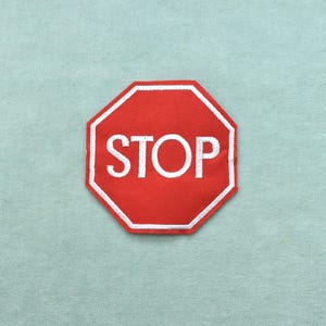 May include: A red, octagonal patch with the word "STOP" in white, outlined in white. The patch is on a light blue fabric background. This patch is suitable for clothing or accessories.