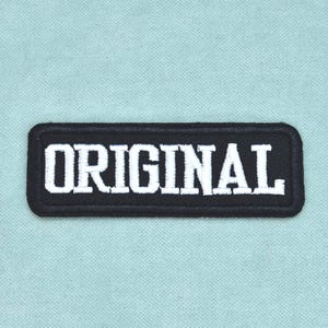 Peut inclure: Un écusson rectangulaire noir brodé aux coins arrondis portant le mot ORIGINAL cousu en lettres capitales blanches et grasses. L'écusson est centré sur un tissu bleu clair texturé avec un motif tissé visible.