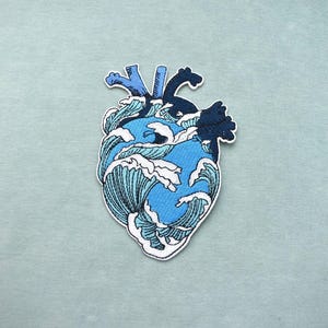 Op de afbeelding: Een geborduurde patch in de vorm van een anatomisch hart, met een ontwerp van beukende oceaangolven in verschillende tinten blauw en wit, met donkerblauwe accenten aan de bovenkant en een witte rand die de hele hartvorm omlijnt.
