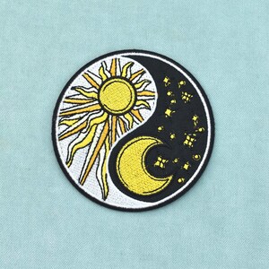 Op de afbeelding: Een ronde geborduurde patch met een yin en yang ontwerp. De ene helft is wit met een gele zon en stralende zonnestralen, terwijl de andere helft zwart is met een gele maansikkel en verschillende kleine gele vierpuntige sterren.