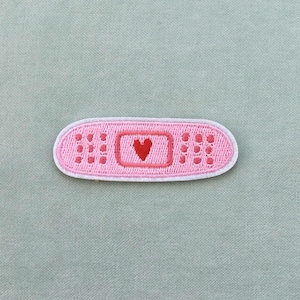 Könnte beinhalten: Ein rosa bestickter Pflaster-Aufnäher mit weißem Rand. Der Aufnäher zeigt ein rotes Herz in der Mitte und gepunktete Details auf jeder Seite. Der Aufnäher ist etwa 7,6 cm lang.