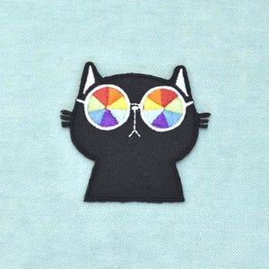 Può includere: Una toppa ricamata raffigurante una testa di gatto nero con orecchie interne e baffi bianchi. Il gatto indossa occhiali rotondi con lenti caratterizzate da un motivo arcobaleno segmentato e multicolore in rosso, arancione, giallo, verde, blu e viola.