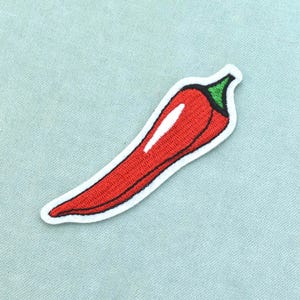 Peut inclure: Un écusson brodé en forme de piment. Le piment est rouge vif avec un reflet blanc sur le côté et une tige verte au sommet. L'ensemble du motif est souligné par une épaisse bordure blanche, créant une forme bien définie et contrastée.