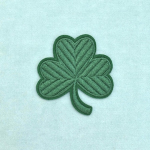 Peut inclure: Un écusson brodé en forme de trèfle vert foncé avec trois feuilles arrondies et une tige incurvée. Le motif est détaillé par des lignes de couture diagonales qui suivent la forme de chaque feuille, créant un aspect texturé sur un tissu bleu clair.