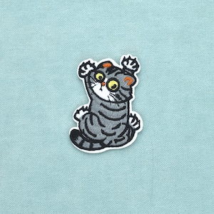 Op de afbeelding: Een geborduurde patch met een grijze cyperse kat met donkergrijze strepen, grote gele ogen en oranje oortoppen. De kat is afgebeeld in een zittende houding met de voorpoten omhoog, waarbij witte klauwen zichtbaar zijn op een witte rand.