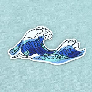 Peut inclure: Un écusson brodé représentant une vague stylisée dans des tons de bleu foncé, bleu moyen et bleu clair avec des crêtes d'écume blanches, bordé d'un contour blanc épais sur un fond en tissu bleu clair uni.