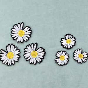 Op de afbeelding: Vijf geborduurde madeliefje-patches met witte bloemblaadjes, gele harten en zwarte omlijningen. De set bevat drie grotere madeliefje-patches en twee kleinere, allemaal met een cirkelvormig ontwerp met gedetailleerd stikwerk.