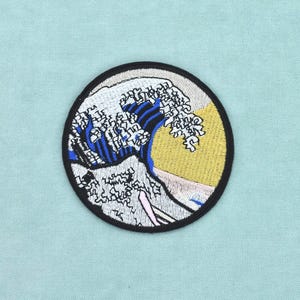 以下が含まれることがあります： 波をモチーフにした円形の刺繍ワッペン。青と白の色調で描かれた大きな波と、背景にある黄色い太陽が特徴です。ワッペン全体は太い黒い縁取りで囲まれています。