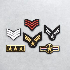 Puede incluir: Colección de parches bordados de insignias de rango militar. Los parches presentan varios diseños, incluyendo galones, estrellas y una estrella dentro de un círculo, en colores negro, dorado, rojo, blanco y azul. Estos parches indican el rango.
