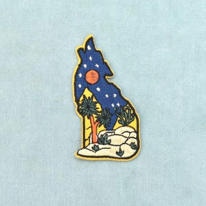 Pode incluir: Um emblema bordado com o formato do estado de Idaho, apresentando a silhueta de um coiote a uivar preenchida com um céu noturno azul-escuro, estrelas brancas e um sol laranja. A parte inferior mostra uma paisagem desértica com árvores verdes.