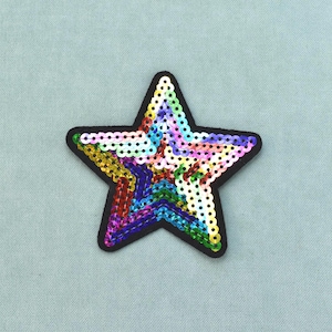 May include: A five-pointed star patch covered in multicolored sequins arranged in a rainbow pattern. The star features a solid black border outlining its shape, with individual sequins reflecting light in shades of red, orange, yellow, green, blue, and purple.