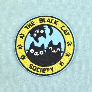 May include: A circular embroidered patch featuring a light blue center with three black cats and a yellow border. The text THE BLACK CAT SOCIETY is stitched in black along the yellow border, which also includes small black paw print icons.